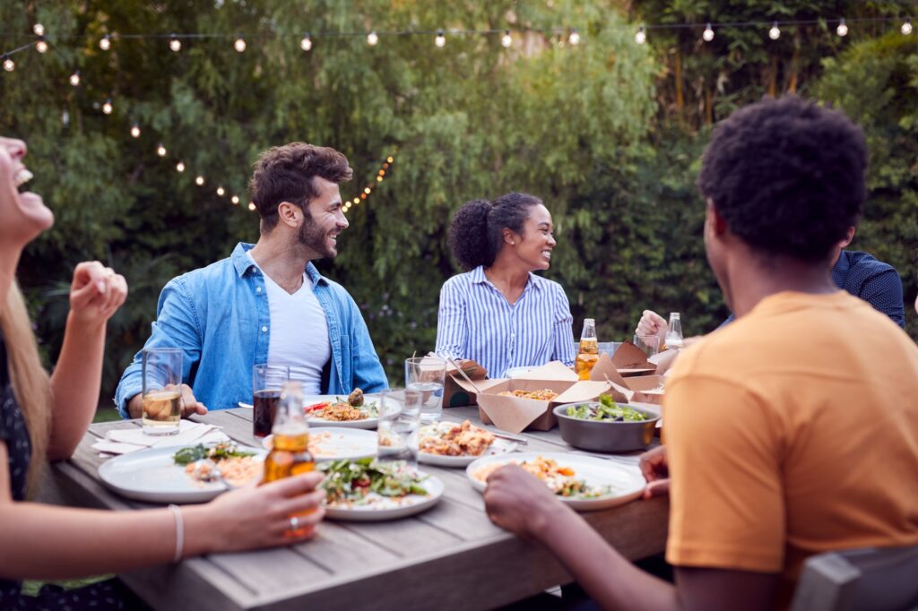 Multi-Cultural Friends At Home Sitting At Table Enjoying Food At Summer Garden Party